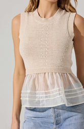 A woman is wearing a sleeveless knit top with a round neckline in a light beige color. The top has a textured, open-knit pattern running vertically down the front, with a ribbed band at the waist. The bottom of the top features a layered, sheer, cream-colored fabric with horizontal stripes, creating a peplum-like effect. She is also wearing light blue denim jeans.